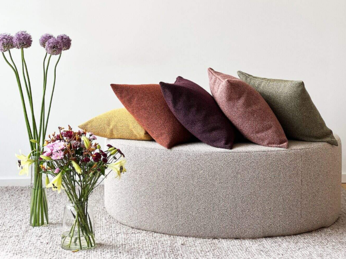 Ein großes rundes Sitzkisten aus grobem grauem Bezugsstoff. Darauf liegen fünf Unikissen in verschiedenen Farben aus dem gleichen Material. Links stehen zwei hohe Blumenvasen mit Blumen.